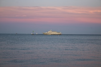 A large yacht is sailing on calm waters under a pastel-colored sky with shades of pink and blue. The horizon is dotted with a couple of small sailboats in the distance, adding depth to the serene seascape.
