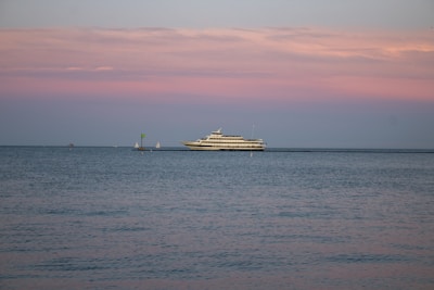 A large yacht is sailing on calm waters under a pastel-colored sky with shades of pink and blue. The horizon is dotted with a couple of small sailboats in the distance, adding depth to the serene seascape.