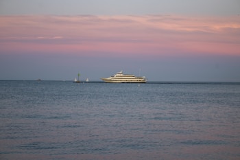 A large yacht is sailing on calm waters under a pastel-colored sky with shades of pink and blue. The horizon is dotted with a couple of small sailboats in the distance, adding depth to the serene seascape.