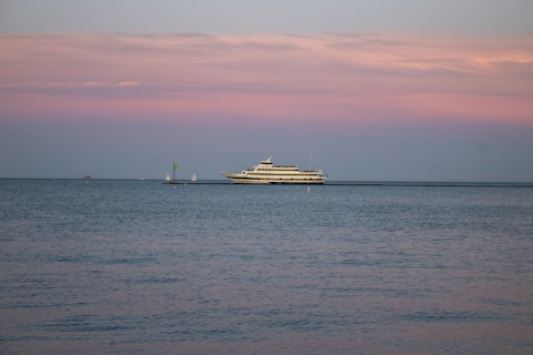 A large yacht is sailing on calm waters under a pastel-colored sky with shades of pink and blue. The horizon is dotted with a couple of small sailboats in the distance, adding depth to the serene seascape.