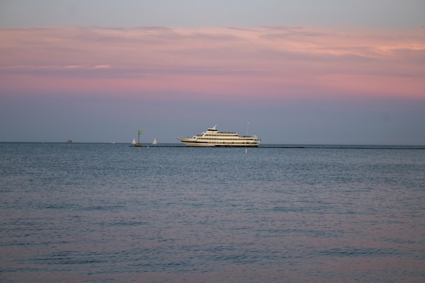 A large yacht is sailing on calm waters under a pastel-colored sky with shades of pink and blue. The horizon is dotted with a couple of small sailboats in the distance, adding depth to the serene seascape.