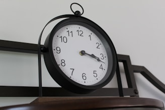 A round wall clock with a black frame, featuring a white face, black numbers, and black hands displaying the time just past three o'clock. It is mounted on a metal structure with a basic, industrial design against a plain background.