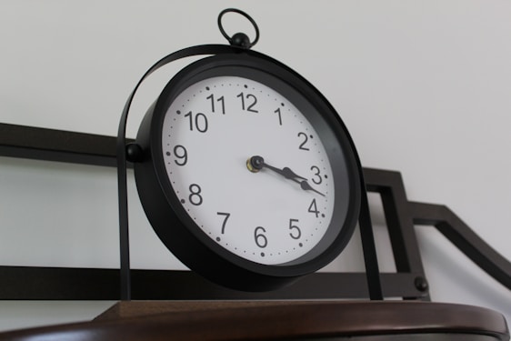A round wall clock with a black frame, featuring a white face, black numbers, and black hands displaying the time just past three o'clock. It is mounted on a metal structure with a basic, industrial design against a plain background.
