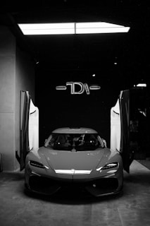 A sleek sports car is parked indoors with its gull-wing doors open. The environment is dimly lit, highlighting the car's aerodynamic design. The background features a large, illuminated logo and minimalistic decor, creating a modern and sophisticated atmosphere.