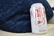 A can of Diet Dr Pepper rests against a textured dark blue fabric background. The can is silver with distinctive red branding and text indicating it is a diet version of the beverage. The perspective suggests the can is tilted slightly toward the viewer.