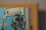 An ophthalmology device, known as a phoropter, is mounted on a mechanical arm against a teal-colored wall. The device has numerous dials and lenses used for vision testing.