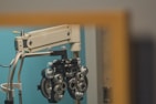 An ophthalmology device, known as a phoropter, is mounted on a mechanical arm against a teal-colored wall. The device has numerous dials and lenses used for vision testing.