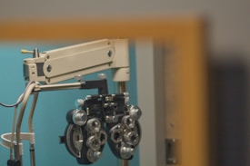 An ophthalmology device, known as a phoropter, is mounted on a mechanical arm against a teal-colored wall. The device has numerous dials and lenses used for vision testing.