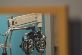 An ophthalmology device, known as a phoropter, is mounted on a mechanical arm against a teal-colored wall. The device has numerous dials and lenses used for vision testing.