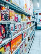 A grocery store aisle filled with a wide variety of cereal boxes neatly arranged on shelves. The cereals include brands like Chocapic, Alpen, Weetabix, and Kellogg's, among others. Price tags are visible on the shelves, indicating the cost of each item, and the floor is tiled and clean. The aisle is well-lit with fluorescent lighting, and a person is seen in the distance browsing.