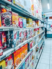A grocery store aisle filled with a wide variety of cereal boxes neatly arranged on shelves. The cereals include brands like Chocapic, Alpen, Weetabix, and Kellogg's, among others. Price tags are visible on the shelves, indicating the cost of each item, and the floor is tiled and clean. The aisle is well-lit with fluorescent lighting, and a person is seen in the distance browsing.