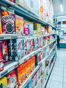 A grocery store aisle filled with a wide variety of cereal boxes neatly arranged on shelves. The cereals include brands like Chocapic, Alpen, Weetabix, and Kellogg's, among others. Price tags are visible on the shelves, indicating the cost of each item, and the floor is tiled and clean. The aisle is well-lit with fluorescent lighting, and a person is seen in the distance browsing.