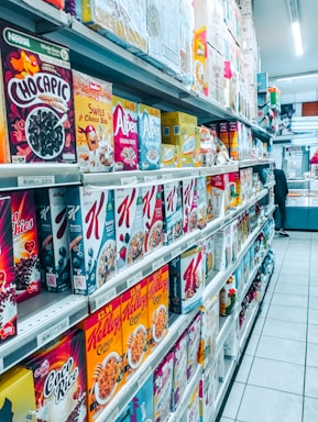 A grocery store aisle filled with a wide variety of cereal boxes neatly arranged on shelves. The cereals include brands like Chocapic, Alpen, Weetabix, and Kellogg's, among others. Price tags are visible on the shelves, indicating the cost of each item, and the floor is tiled and clean. The aisle is well-lit with fluorescent lighting, and a person is seen in the distance browsing.