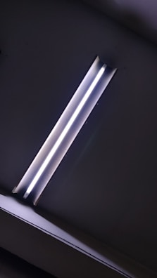 A fluorescent light fixture mounted on a ceiling, emitting a bright white glow. The surroundings are mostly dark, highlighting the light as the focal point.