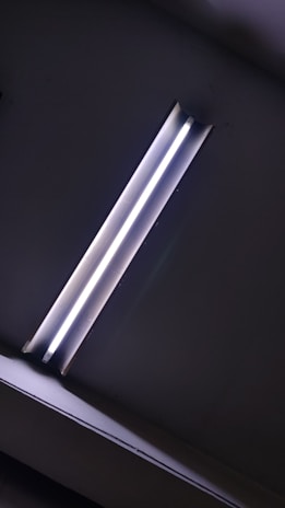 A fluorescent light fixture mounted on a ceiling, emitting a bright white glow. The surroundings are mostly dark, highlighting the light as the focal point.