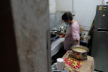 A person stands in a small kitchen next to a counter that holds various cooking ingredients and utensils. The person is wearing a pink checkered shirt and a face mask, focused on preparing something on a portable stove. There is a large pot on the counter containing a creamy mixture, next to a collection of colorful round objects, possibly snacks or ingredients. The kitchen appears cluttered with various items, including bottles and cooking pots.