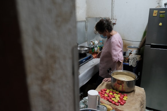 A person stands in a small kitchen next to a counter that holds various cooking ingredients and utensils. The person is wearing a pink checkered shirt and a face mask, focused on preparing something on a portable stove. There is a large pot on the counter containing a creamy mixture, next to a collection of colorful round objects, possibly snacks or ingredients. The kitchen appears cluttered with various items, including bottles and cooking pots.
