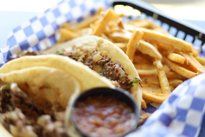 A plate with two tacos filled with seasoned meat and topped with diced onions and herbs is placed in a checkered paper-lined basket. Next to the tacos are crispy golden French fries. There is a small container of salsa, likely used as a condiment for the tacos.