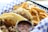 A plate with two tacos filled with seasoned meat and topped with diced onions and herbs is placed in a checkered paper-lined basket. Next to the tacos are crispy golden French fries. There is a small container of salsa, likely used as a condiment for the tacos.