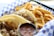 A plate with two tacos filled with seasoned meat and topped with diced onions and herbs is placed in a checkered paper-lined basket. Next to the tacos are crispy golden French fries. There is a small container of salsa, likely used as a condiment for the tacos.