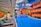 A modern pool setting surrounded by vibrant orange buildings under a bright blue sky with scattered clouds. The pool has a distinctive angular shape with wooden lounge chairs arranged under a pergola. Lush greenery and palm trees accentuate the scene, adding a tropical feel.