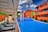 A modern pool setting surrounded by vibrant orange buildings under a bright blue sky with scattered clouds. The pool has a distinctive angular shape with wooden lounge chairs arranged under a pergola. Lush greenery and palm trees accentuate the scene, adding a tropical feel.
