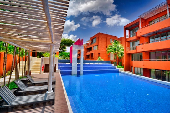 A modern pool setting surrounded by vibrant orange buildings under a bright blue sky with scattered clouds. The pool has a distinctive angular shape with wooden lounge chairs arranged under a pergola. Lush greenery and palm trees accentuate the scene, adding a tropical feel.