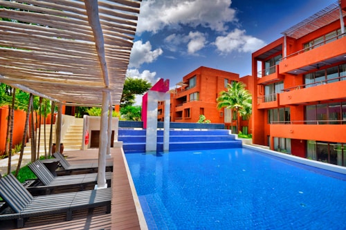 A modern pool setting surrounded by vibrant orange buildings under a bright blue sky with scattered clouds. The pool has a distinctive angular shape with wooden lounge chairs arranged under a pergola. Lush greenery and palm trees accentuate the scene, adding a tropical feel.