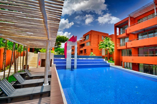 A modern pool setting surrounded by vibrant orange buildings under a bright blue sky with scattered clouds. The pool has a distinctive angular shape with wooden lounge chairs arranged under a pergola. Lush greenery and palm trees accentuate the scene, adding a tropical feel.