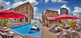 A luxurious rooftop setting featuring a swimming pool with cabanas and white pool loungers. The area is surrounded by elegant architecture with classical design elements, including large stone buildings. Bright red umbrellas provide shade for the modern outdoor furniture on the wooden deck, where a few tables and chairs are placed for relaxation. The sky is clear with a few fluffy clouds, enhancing the vibrant atmosphere.