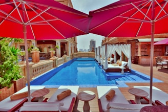 Luxury rooftop pool area with elegant loungers and umbrellas providing shade. The pool features clear blue water and is complemented by canopied daybeds adorned with cushions. Architectural elements in earth tones along with greenery create a serene and opulent atmosphere. A cityscape is visible in the distance.