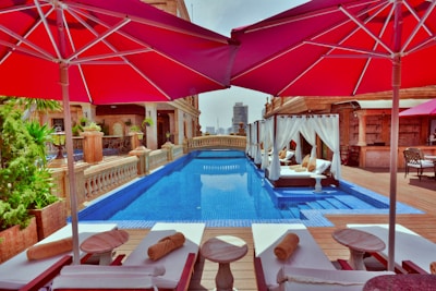 Luxury rooftop pool area with elegant loungers and umbrellas providing shade. The pool features clear blue water and is complemented by canopied daybeds adorned with cushions. Architectural elements in earth tones along with greenery create a serene and opulent atmosphere. A cityscape is visible in the distance.