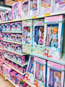 A store shelf filled with various boxed dolls and toy sets. The packaging features colorful designs and illustrations, with several dolls in different outfits visible through transparent windows. Brands like 'Little Lacee' and 'Unity Girl' are prominently displayed, offering themed sets such as baby dolls and doctor kits.