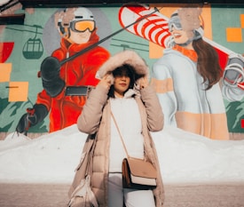 A person in a winter coat with a fur-lined hood stands in front of a colorful mural featuring large images of skiers. The mural includes vibrant shades of red, orange, and green, and depicts a ski resort theme with skiers wearing helmets and goggles. The snowy ground suggests a cold environment.