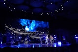 A stage performance with musicians playing under dramatic lighting. A large screen displays a fictional scene with a creature on a futuristic ship against a cosmic blue background. Musical equipment and instruments are visible.