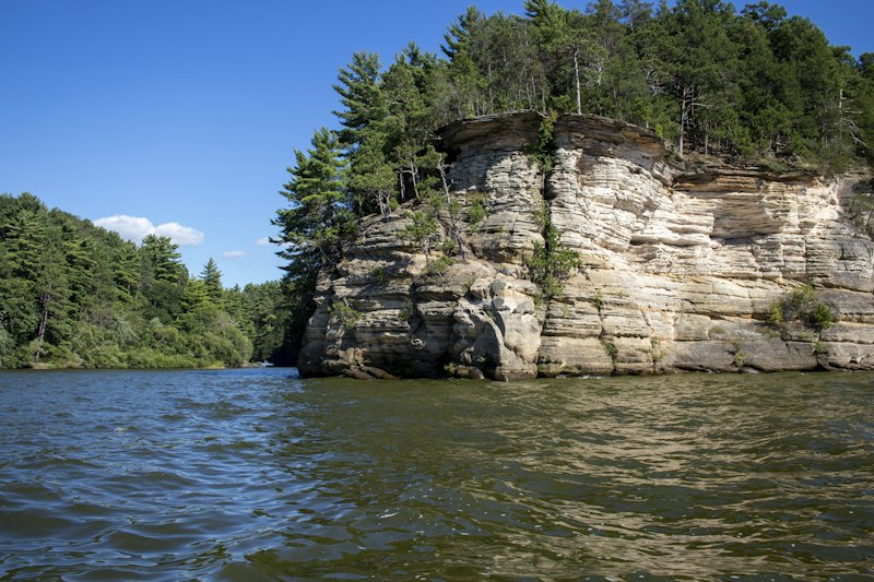 Pine-topped sandstone cliff rising straight out of the Wisconsin River under a deep blue sky in the Upper Dells