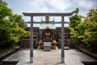 A traditional Japanese Shinto shrine is framed by a wooden torii gate. The shrine is surrounded by lush green foliage and a stone wall backdrop, adding a serene and natural setting to the scene. The ground appears to be made of stone tiles, leading up to the shrine.