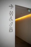 A corridor entrance with several signs on the wall indicating directions for restrooms, accessible facilities, baby changing area, and luggage storage. The signs are mounted on a light-colored wall, and an illuminated exit sign is visible above the corridor.