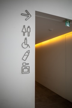 A corridor entrance with several signs on the wall indicating directions for restrooms, accessible facilities, baby changing area, and luggage storage. The signs are mounted on a light-colored wall, and an illuminated exit sign is visible above the corridor.