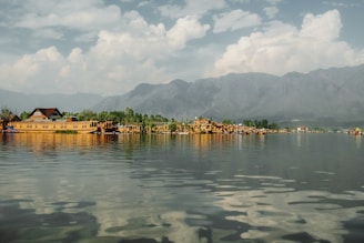 A scenic view of houseboats floating on a calm lake, with lush greenery and trees along the shore. Majestic mountains form the backdrop under a partly cloudy sky, reflecting in the clear water.