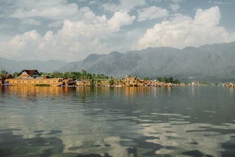 A scenic view of houseboats floating on a calm lake, with lush greenery and trees along the shore. Majestic mountains form the backdrop under a partly cloudy sky, reflecting in the clear water.