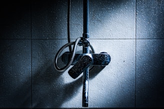 A dimly lit bathroom wall with a metallic shower fixture and hose. The tiles have a textured appearance, and subtle lighting creates a dramatic contrast with shadows.