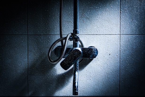 A dimly lit bathroom wall with a metallic shower fixture and hose. The tiles have a textured appearance, and subtle lighting creates a dramatic contrast with shadows.
