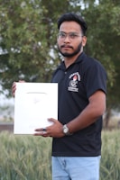 A man wearing a black polo shirt and glasses holds a large silver plaque with a play button symbol. He stands outdoors in front of a lush green backdrop.