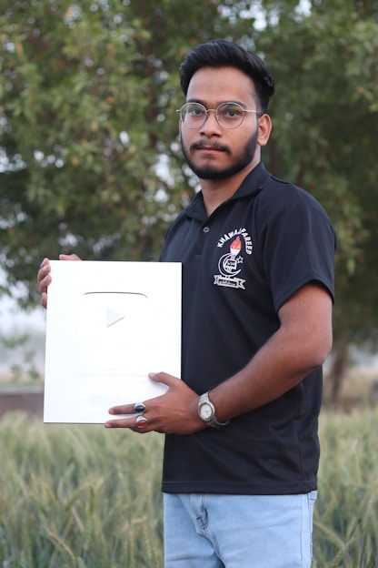 A man wearing a black polo shirt and glasses holds a large silver plaque with a play button symbol. He stands outdoors in front of a lush green backdrop.