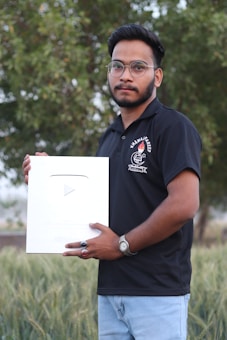 A man wearing a black polo shirt and glasses holds a large silver plaque with a play button symbol. He stands outdoors in front of a lush green backdrop.