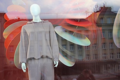 A mannequin dressed in a casual gray hoodie and pants is displayed in a store window, with colorful abstract shapes in red, orange, and blue hanging behind it. The glass reflects an urban scene of buildings, adding a layered and artistic effect.