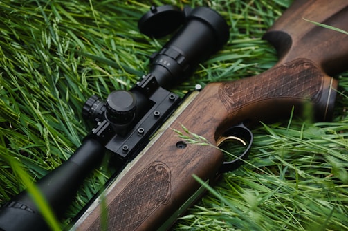 A rifle with a wooden stock and a mounted scope lies on a bed of lush green grass. The detailed wood engraving on the stock is visible, and the black metallic parts of the rifle contrast sharply with the greenery.