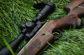 A rifle with a wooden stock and a mounted scope lies on a bed of lush green grass. The detailed wood engraving on the stock is visible, and the black metallic parts of the rifle contrast sharply with the greenery.