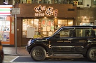 A street view featuring the front of a restaurant with bright signage written in a combination of English and what appears to be Japanese characters. There is a large black SUV parked in front of the restaurant. The restaurant windows display various posters and advertisements, adding to the vibrant and busy atmosphere. Adjacent to the restaurant, a brightly lit storefront with additional posters is visible.